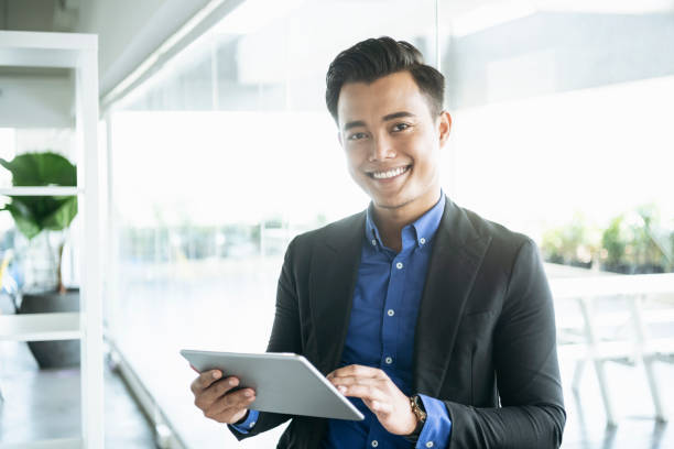 Man in his 20s smiling towards camera and using tablet in modern office